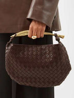 Person in a brown leather jacket holding Noémie brown woven vegan leather handbag with gold-tone handle