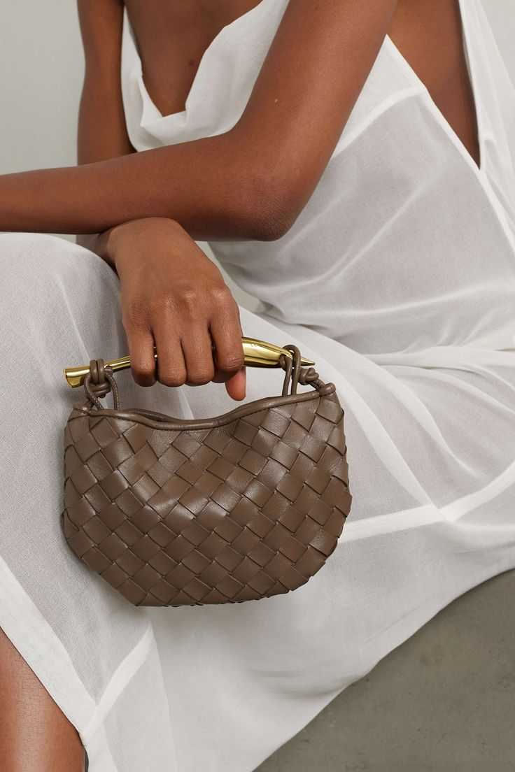 Person in a white dress holding Noémie brown woven vegan leather handbag with gold-tone hardware