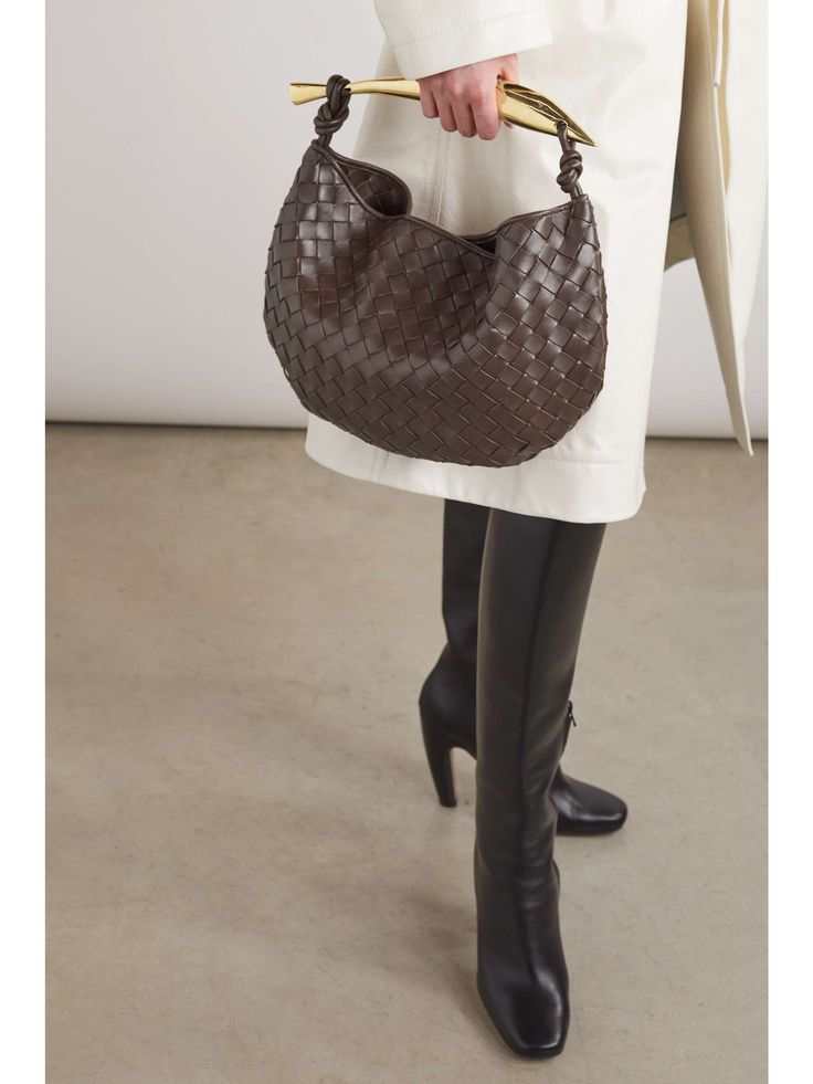 Person in a white coat and black boots holding Noémie brown woven vegan leather handbag with gold-tone hardware