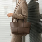 Model carrying Aurelia brown genuine leather tote bag with a stylish woven design, dressed in a plaid suit and standing in front of large windows