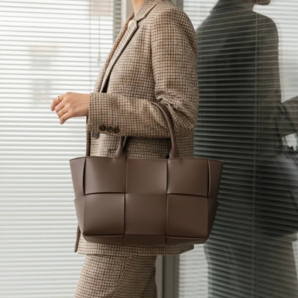 Model carrying Aurelia brown genuine leather tote bag with a stylish woven design, dressed in a plaid suit and standing in front of large windows