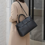 Side view of a woman carrying a sleek black genuine leather tote bag with adjustable shoulder straps, paired with a beige coat