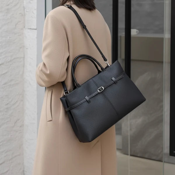 Side view of a woman carrying a sleek black genuine leather tote bag with adjustable shoulder straps, paired with a beige coat