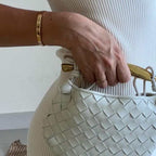 Woman in white holding Noémie cream woven vegan leather handbag with gold-tone hardware
