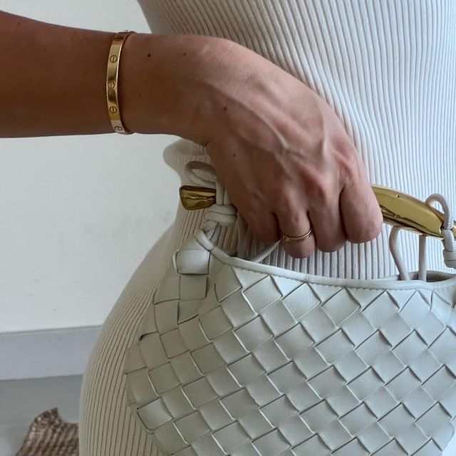 Woman in white holding Noémie cream woven vegan leather handbag with gold-tone hardware