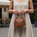 Woman holding a Brown Izelia top grain leather crossbody bag with gold accents in an outdoor setting