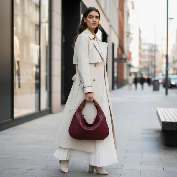 Model standing in an urban setting with a wine red woven vegan leather hobo bag, wearing a beige trench coat